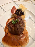 A plated gourmet steak dish with a tender braised beef cheek, rich orange-brown gravy sauce, a potato waffle chip, a mushroom cap, and a garnish of fresh parsley on a white gold-rimmed plate. 