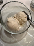 Two scoops of vanilla ice cream in a clear glass bowl with a metal spoon, set on a white plate with a visible watermark overlay.