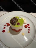 A small pistachio and strawberry mouse cake on a white plate with a gold rim, topped with green mousse, chocolate drizzle, a small cream dollop, and strawberry pieces, photographed in a restaurant setting.