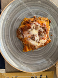 A top-down view of a baked pasta dish, likely lasagna or carbonara, topped with melted white cheese and browned sausage, served on a round gray ceramic plate.