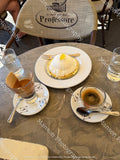 A table setting in a cafe featuring a plate of dessert topped with a yellow lemon curd, alongside espresso and iced coffee drinks on white saucers, with a chair in the background.