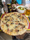 High-angle view of a whole Italian white pizza topped with sliced mushrooms, crumbled sausage, and melted cheese. Served at an outdoor restaurant table alongside a side salad, Italian beer, and lemon soda.