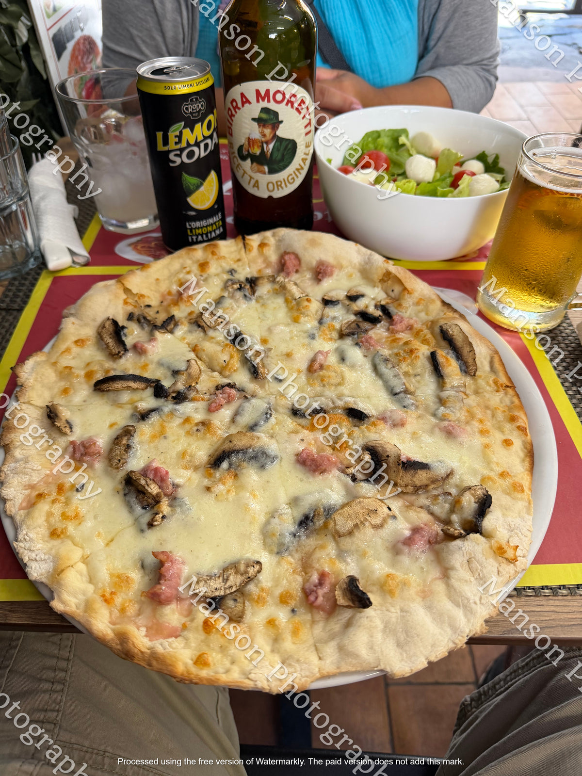 High-angle view of a whole Italian white pizza topped with sliced mushrooms, crumbled sausage, and melted cheese. Served at an outdoor restaurant table alongside a side salad, Italian beer, and lemon soda.