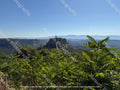 A scenic landscape photograph of the historic hilltop village of Civita di Bagnoregio. A stone village perched on a rocky hilltop above a lush green valley, with layered blue mountain ranges in the distance under a clear blue sky. 