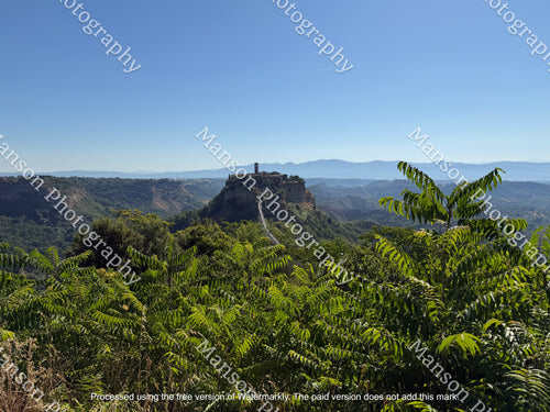 A scenic landscape photograph of the historic hilltop village of Civita di Bagnoregio. A stone village perched on a rocky hilltop above a lush green valley, with layered blue mountain ranges in the distance under a clear blue sky. 