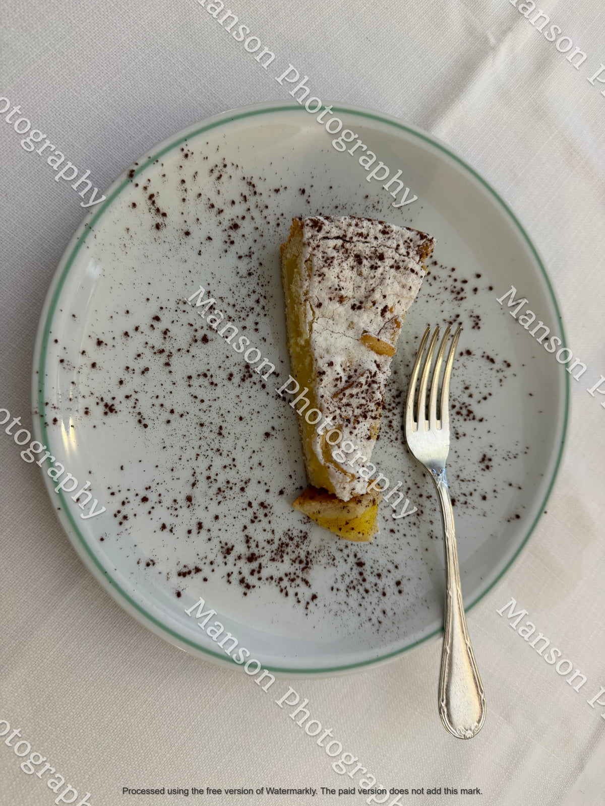 An overhead shot of a slice of dessert cake topped with powdered sugar, served alongside a dessert fork on a white porcelain plate. The plate is dusted with fine cocoa powder and set against a neutral, textured white tablecloth.
