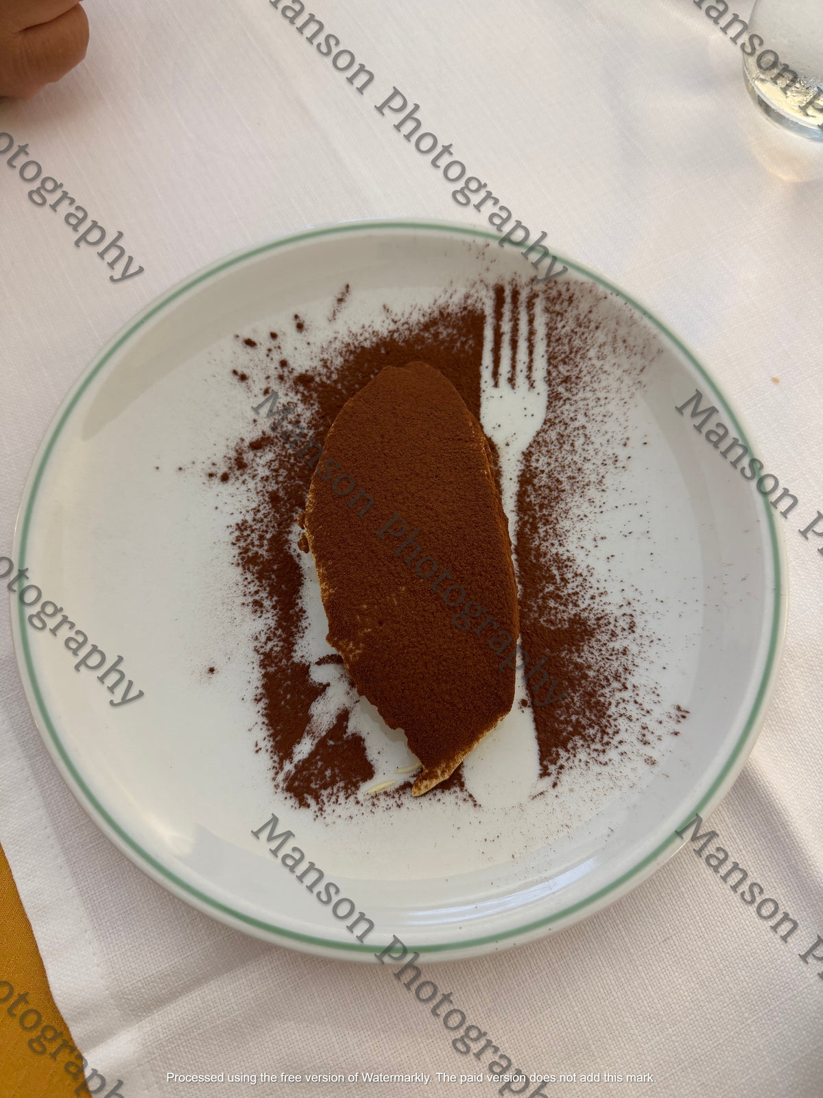 An overhead view of a portion of tiramisu set on a white ceramic plate dusted with cocoa powder. The plating features a negative space fork stencil pattern left in the cocoa dusting, served over a clean white tablecloth.