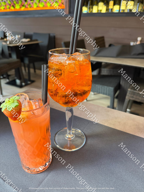 An eye-level shot of two iced cocktails served on a dark tabletop inside a restaurant. Features a classic bright orange Aperol Spritz in a wine glass alongside a pink highball cocktail garnished with a fresh strawberry, both presented with dark straws.