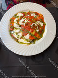 An overhead view of a freshly plated ravioli dish coated in a rich tomato sauce and garnished with melted cream cheese and a green pesto drizzle. Served on a white ceramic plate with a spiraled rim over a dark table setting.
