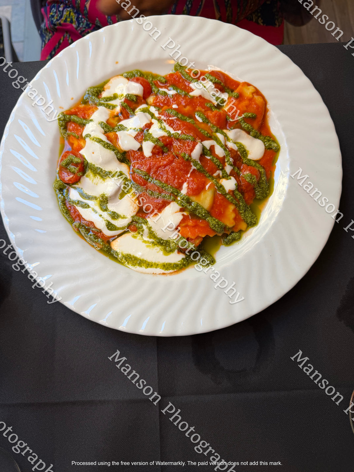 An overhead view of a freshly plated ravioli dish coated in a rich tomato sauce and garnished with melted cream cheese and a green pesto drizzle. Served on a white ceramic plate with a spiraled rim over a dark table setting.