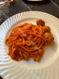 An overhead shot of long tagliatelle pasta coated in vibrant tomato sauce and served alongside three cooked meatballs. Plated on a white ceramic dish with a spiral-patterned rim against a dark tablecloth setting.