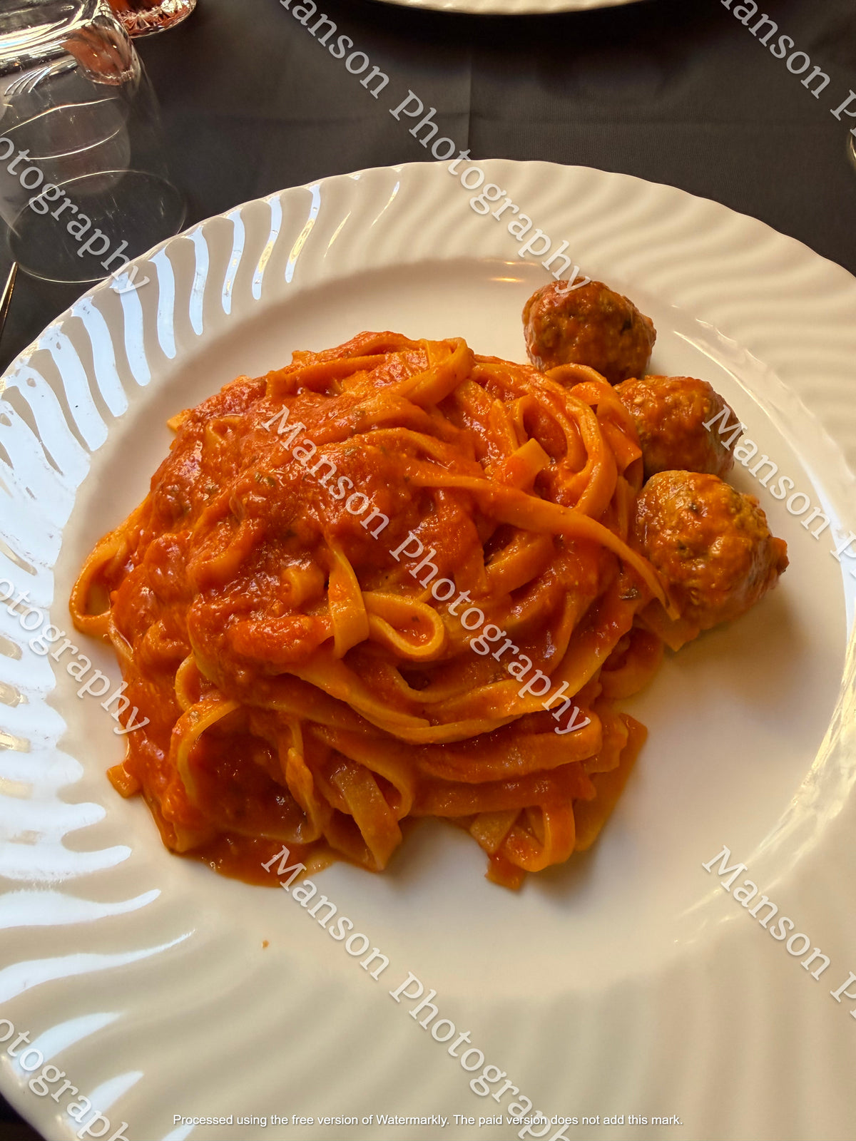 An overhead shot of long tagliatelle pasta coated in vibrant tomato sauce and served alongside three cooked meatballs. Plated on a white ceramic dish with a spiral-patterned rim against a dark tablecloth setting.