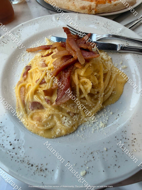 A plate of creamy pasta with crispy bacon pieces on top, served with a fork and knife on a white plate. 